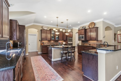 This spacious kitchen features dark wood cabinetry, a large central island with a gas cooktop, and elegant pendant lighting. The open layout flows into a dining area with large windows, providing natural light. The warm hardwood floors and ample counter space make it perfect for entertaining.