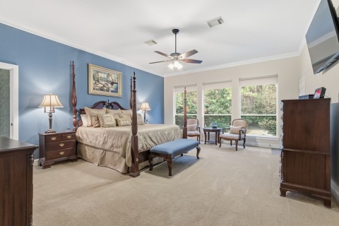 Luxurious primary bedroom featuring a cozy seating area, elegant crown molding, ceiling fan, plush carpet, and stunning backyard views. Automatic blinds are also featured in the bedroom.