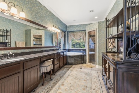 This spacious bathroom features elegant dark wood cabinetry, a long granite countertop with dual sinks, and warm lighting. It includes a luxurious corner bathtub with a picturesque window view and a separate glass-enclosed walk in shower.