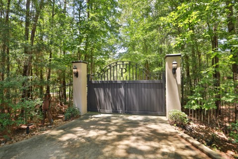 This photo shows a gated driveway entrance surrounded by tall trees, with elegant pillars featuring decorative lanterns. The paved path offers privacy and a welcoming approach to the property.