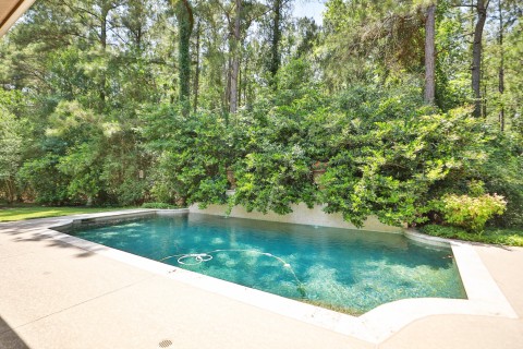 This photo features a serene backyard with a pool surrounded by lush greenery and tall trees, offering a private and tranquil setting. The pool area is bordered by a Sundek surface and has a pebble tech finish. Perfect place for relaxation and your outdoor enjoyment.