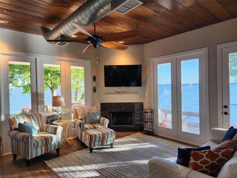 This is a cozy lakeside living room featuring a warm wood ceiling, a fireplace, comfortable seating, and large French doors that offer stunning views of the lake.