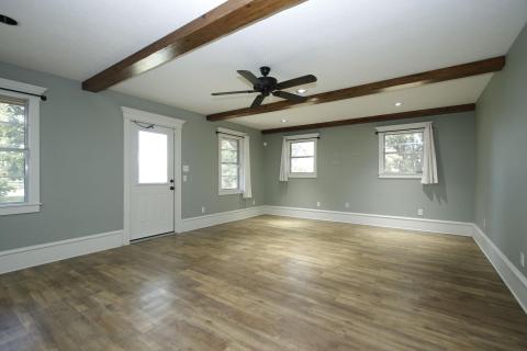 Spacious living area with laminate wood floors, ceiling beams, and a ceiling fan. It features three windows and a door for natural light, with neutral wall colors creating a bright, inviting atmosphere. Spacious living area with laminate wood floors, ceiling beams, and a ceiling fan. It features three windows and a door for natural light, with neutral wall colors creating a bright, inviting atmosphere.