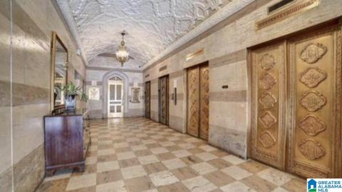 Ornate hallway featuring a vaulted, textured ceiling with decorative detailing, checkered tile flooring, and banded stone walls