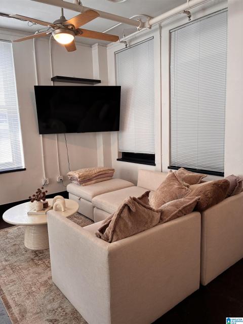 Living area featuring multiple windows with blinds, a ceiling fan with integrated lighting, and exposed ceiling pipes