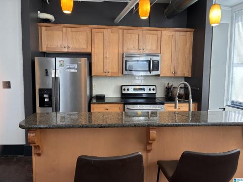 Open-concept kitchen featuring light wood cabinetry, stainless steel appliances, granite countertops, a subway tile backsplash, and a breakfast bar with decorative corbels