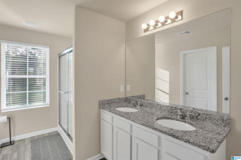 Spa-inspired master bathroom featuring dual vanities with granite countertops, an expansive framed mirror with integrated lighting, a walk-in shower with glass enclosure, and neutral finishes throughout