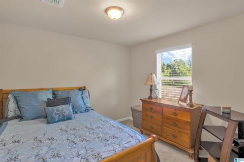 Bright secondary bedroom featuring wood-frame bed, blue patterned bedding, wood dresser with brass hardware, desk workspace, and large window with exterior railing visible